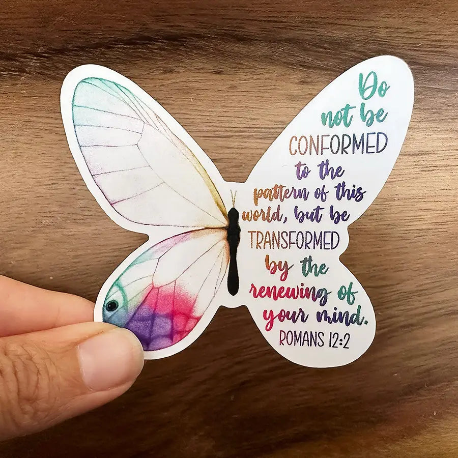 Sticker of a butterfly held in a hand against wood, one half featuring delicate white wings with watercolor pink, purple, and teal, the other half displaying a gradient rainbow quote of Romans 12:2