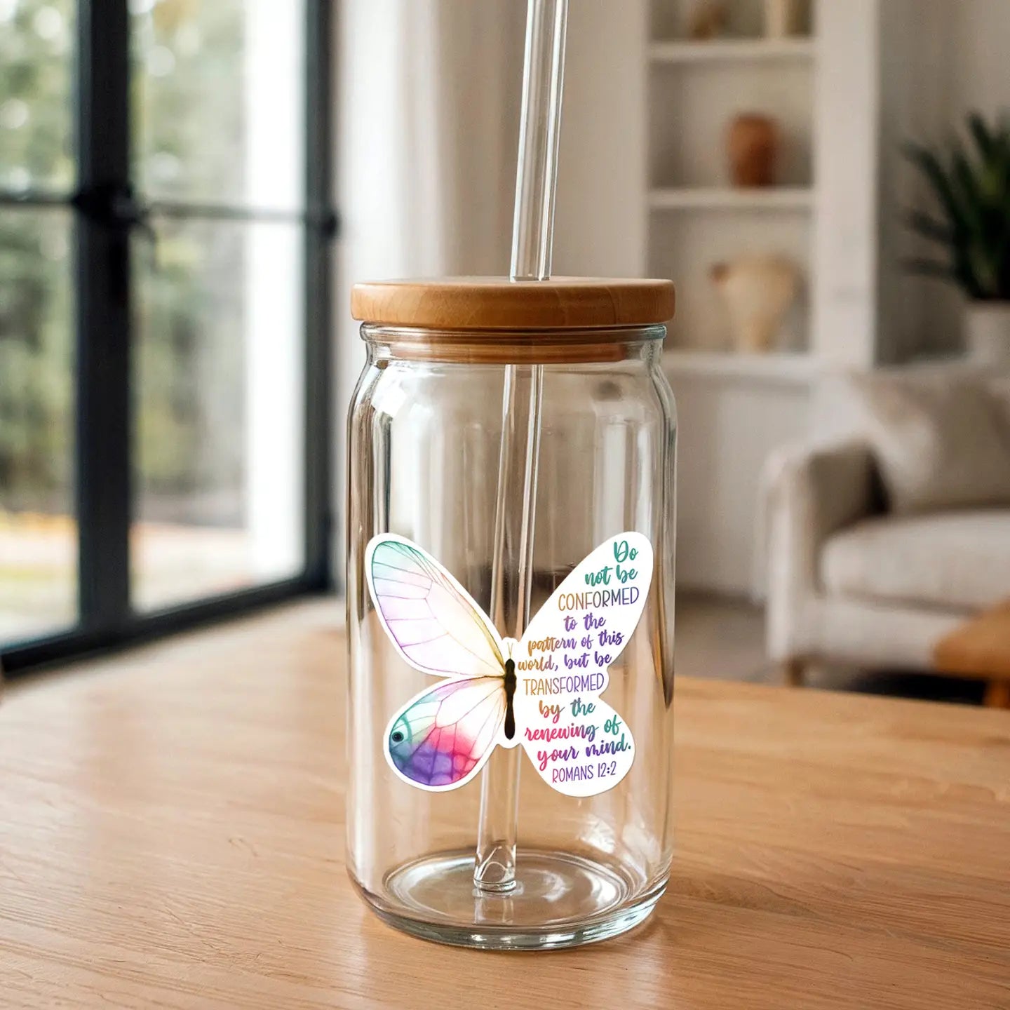 A clear glass tumbler with a bamboo lid and glass straw, adorned with a vibrant watercolor butterfly sticker displaying pastel pink, purple, blue, and green hues and the Romans 12:2 scripture, set on a light wooden table indoors against a soft-focus background of a window and bookshelf