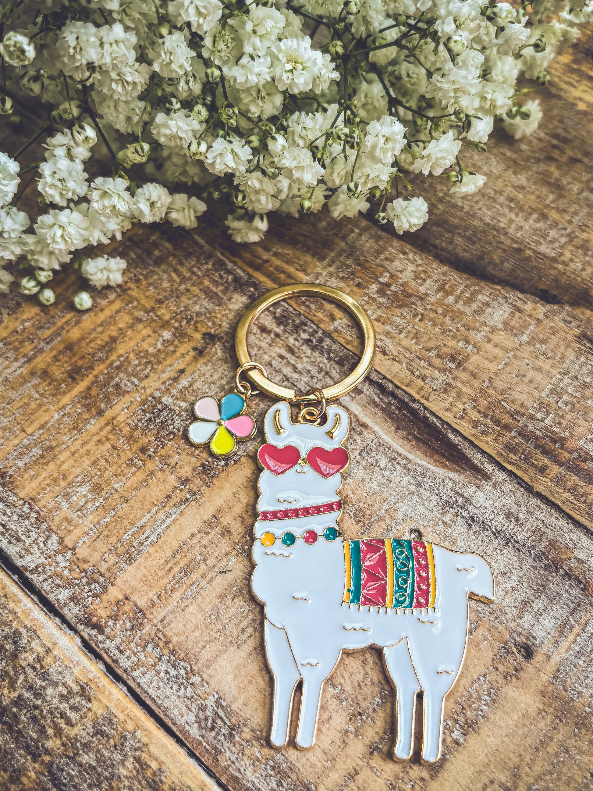 A white llama keychain wearing red heart sunglasses and a colorful patterned blanket on a rustic wooden surface, beside delicate white baby's breath flowers