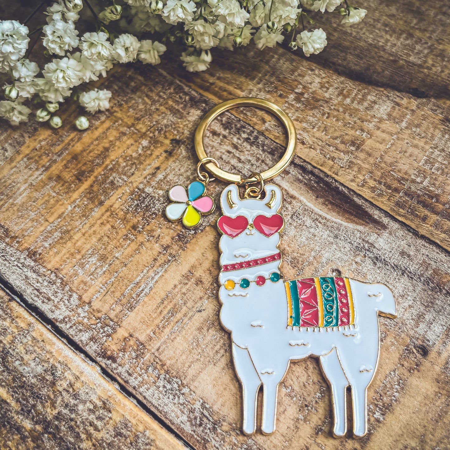 Photograph of a white enamel llama keychain wearing red heart sunglasses and a colorful striped blanket, featuring an attached four-petal flower charm, resting on a rustic wooden surface with white baby's breath flowers in the upper corner