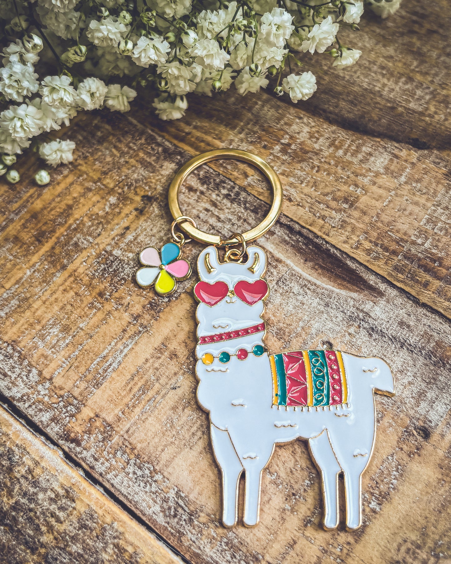 Photograph of a white enamel llama keychain wearing red heart sunglasses and a colorful patterned blanket, with a small multi-colored flower charm on a gold keyring, positioned on a rustic wooden surface alongside white baby's breath flowers
