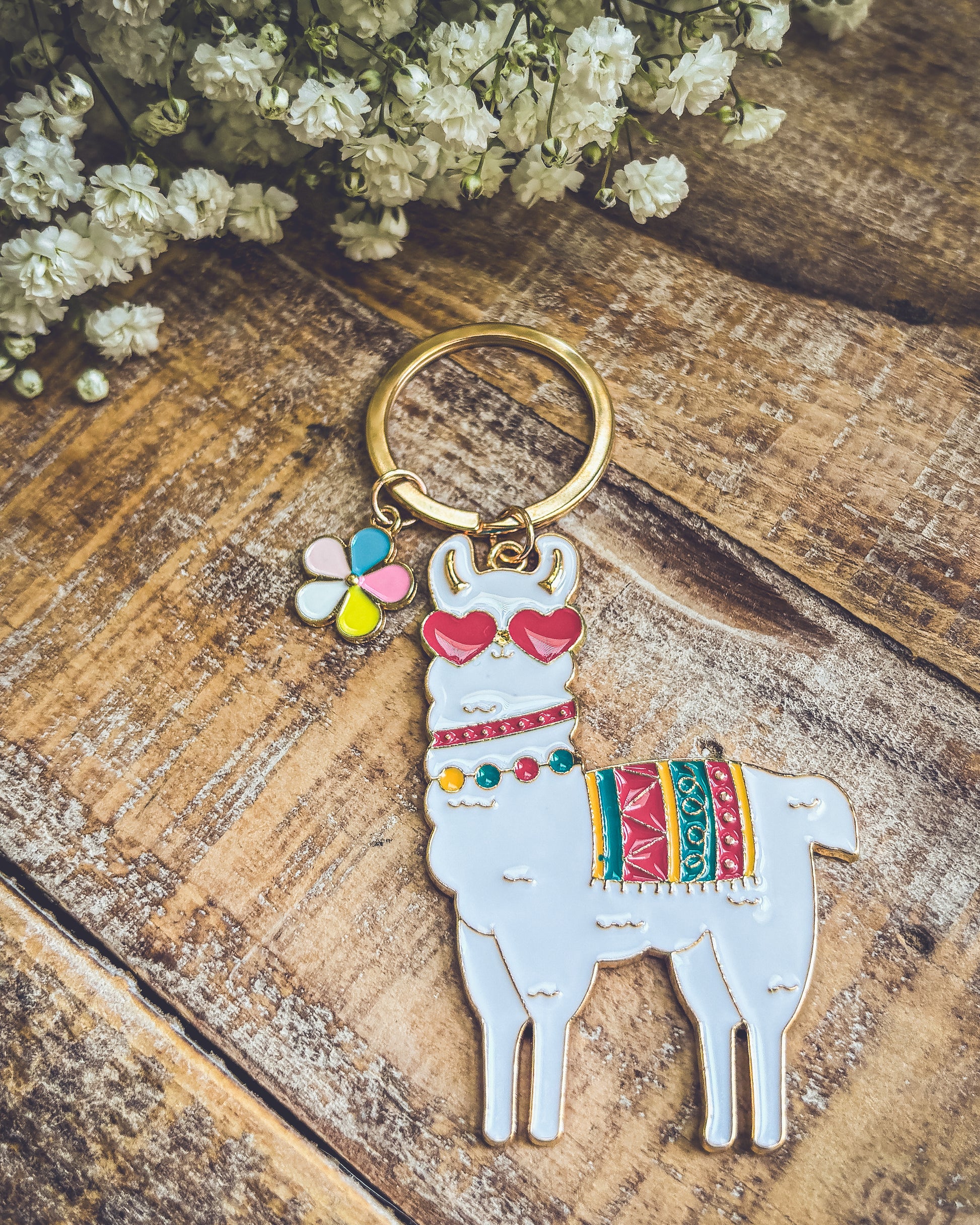 Photograph of a white enamel llama keychain wearing red heart sunglasses and a colorful patterned blanket, with a small multi-colored flower charm on a gold keyring, positioned on a rustic wooden surface alongside white baby's breath flowers