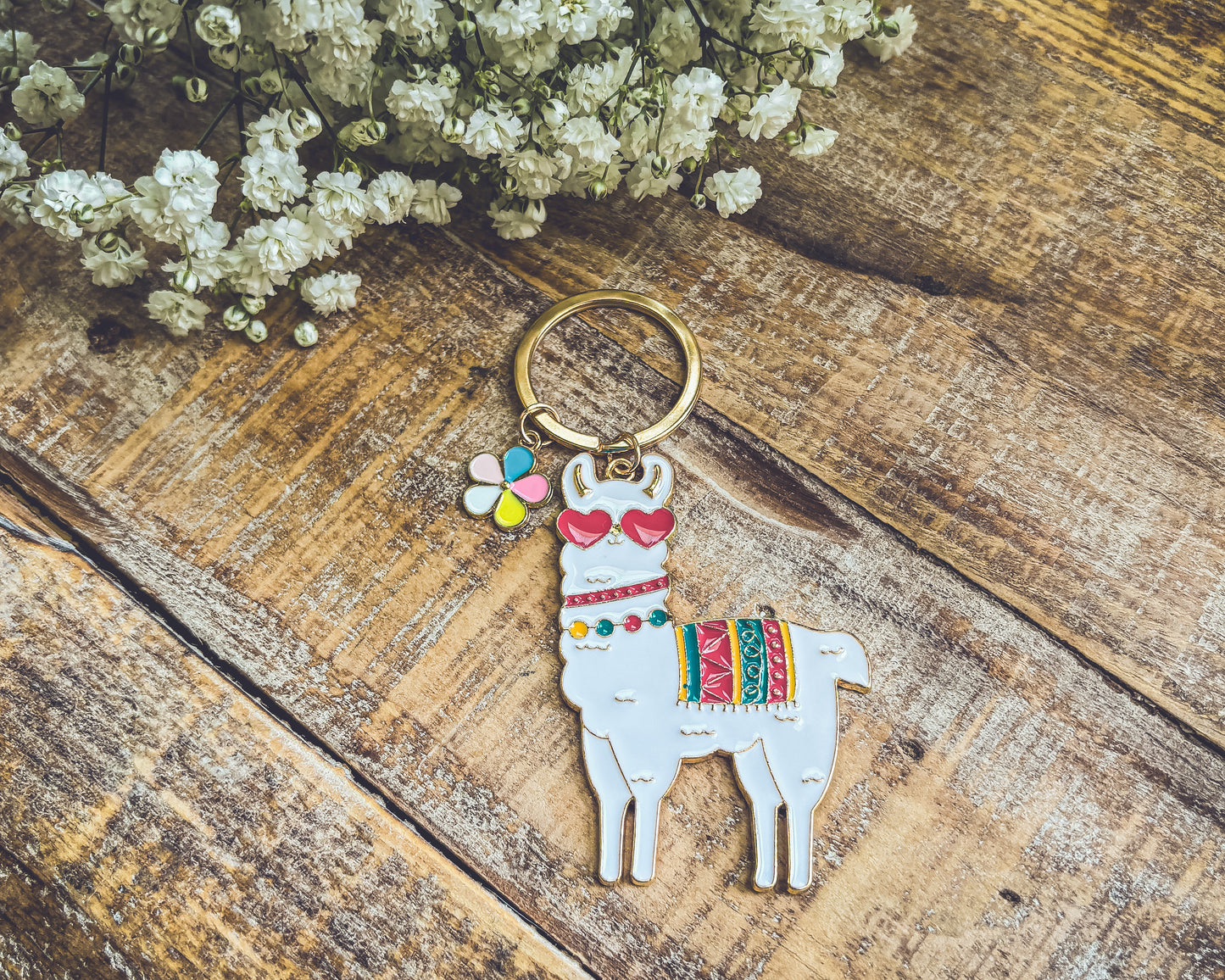 White enamel llama keychain wearing red heart sunglasses with a colorful patterned blanket and a multicolored flower charm resting on rustic wooden planks beside delicate white baby's breath flowers