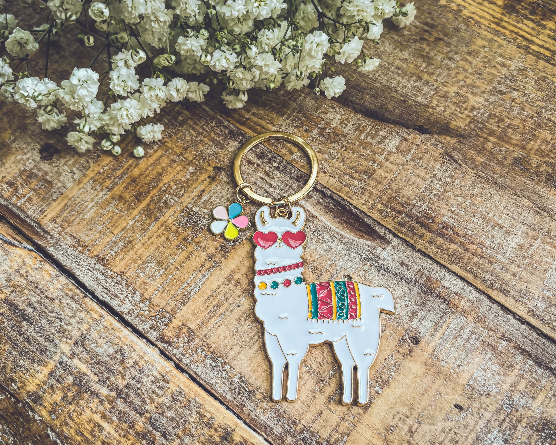 White enamel llama keychain wearing red heart sunglasses with a colorful patterned blanket and a multicolored flower charm resting on rustic wooden planks beside delicate white baby's breath flowers