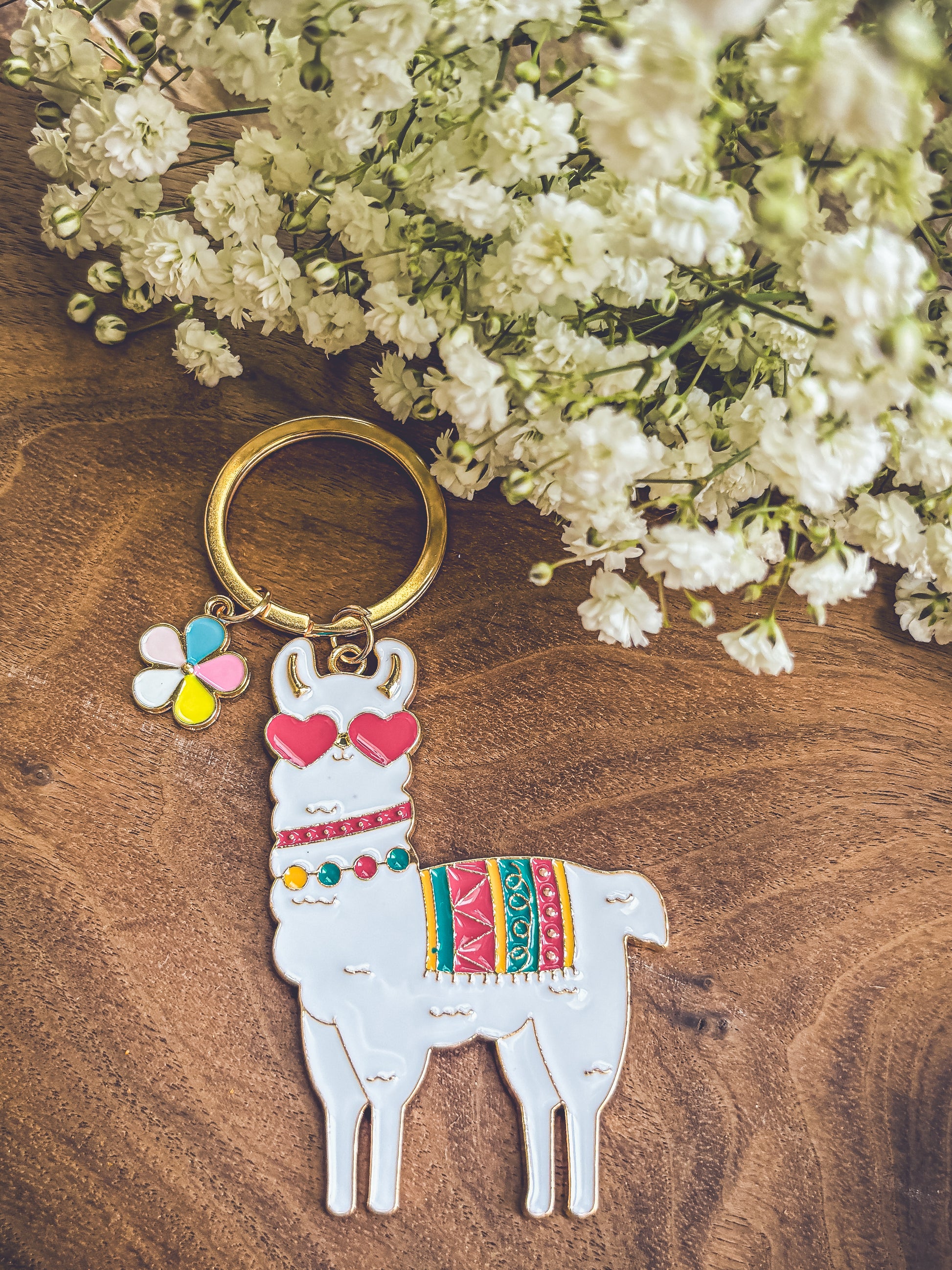 Photograph of a playful white enamel llama keychain featuring red heart-shaped glasses and a vibrant patterned blanket, accompanied by a multicolored flower charm, all resting on a dark rustic wooden surface with delicate white baby's breath flowers