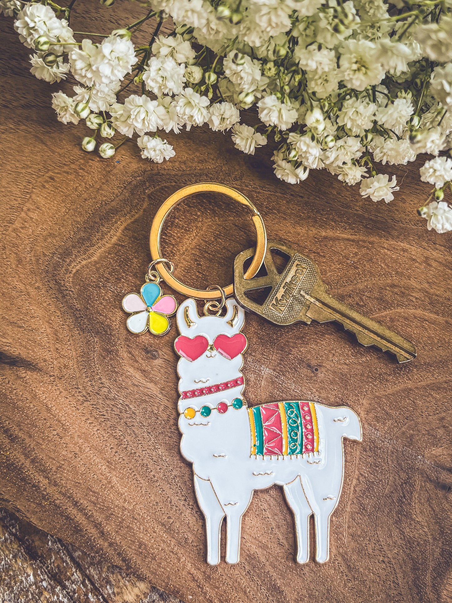 Photograph of a charming white enamel llama keychain wearing pink heart sunglasses and a vibrant striped blanket, accompanied by a bronze key and a multicolor flower charm, all arranged on a warm wooden surface with delicate white baby's breath flowers