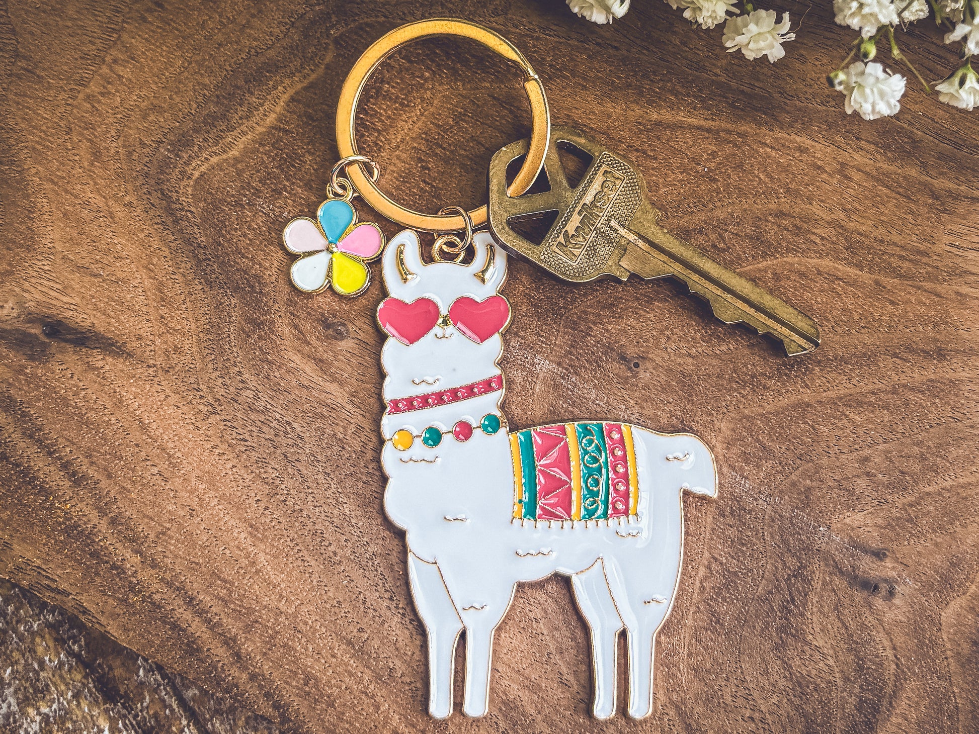 Product photograph showcasing a white enamel llama keychain wearing pink heart sunglasses and a colorful striped blanket, paired with a small multi-color flower charm and a brass key, resting on a textured wooden background with white baby's breath accents