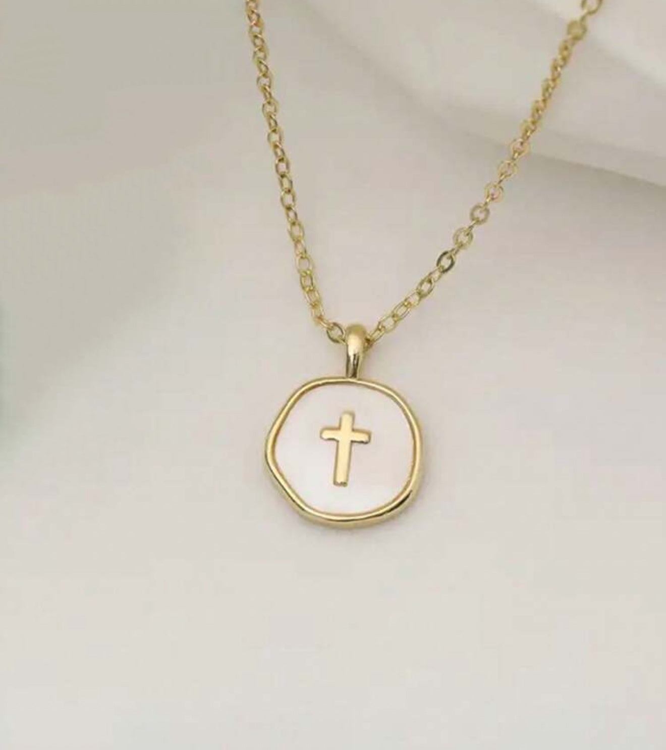 Gold necklace featuring a delicate chain and an irregular white mother-of-pearl round pendant with a raised gold cross