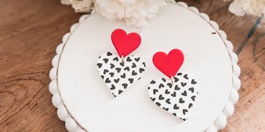 Handmade polymer clay dangle earrings featuring bright red top hearts and white bottom hearts patterned with numerous small black hearts, presented on a white scalloped stand amidst soft white floral accents and a rustic wood table surface