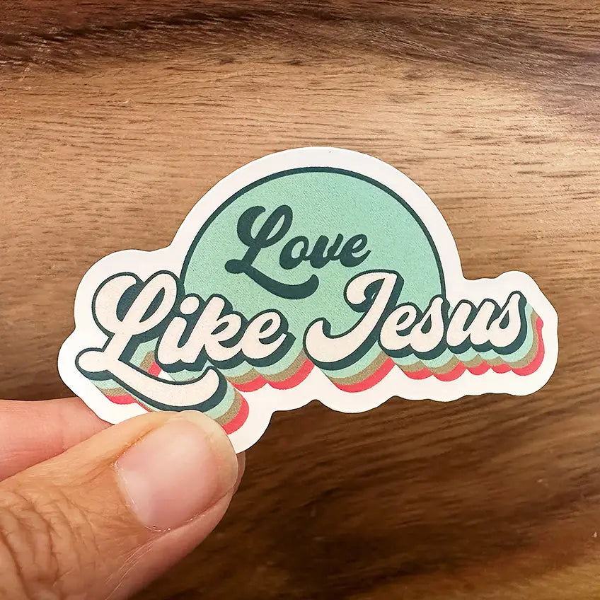 Sticker held by a hand against a wooden background featuring the phrase Love Like Jesus in retro-style teal and cream lettering with red and brown shadow effects