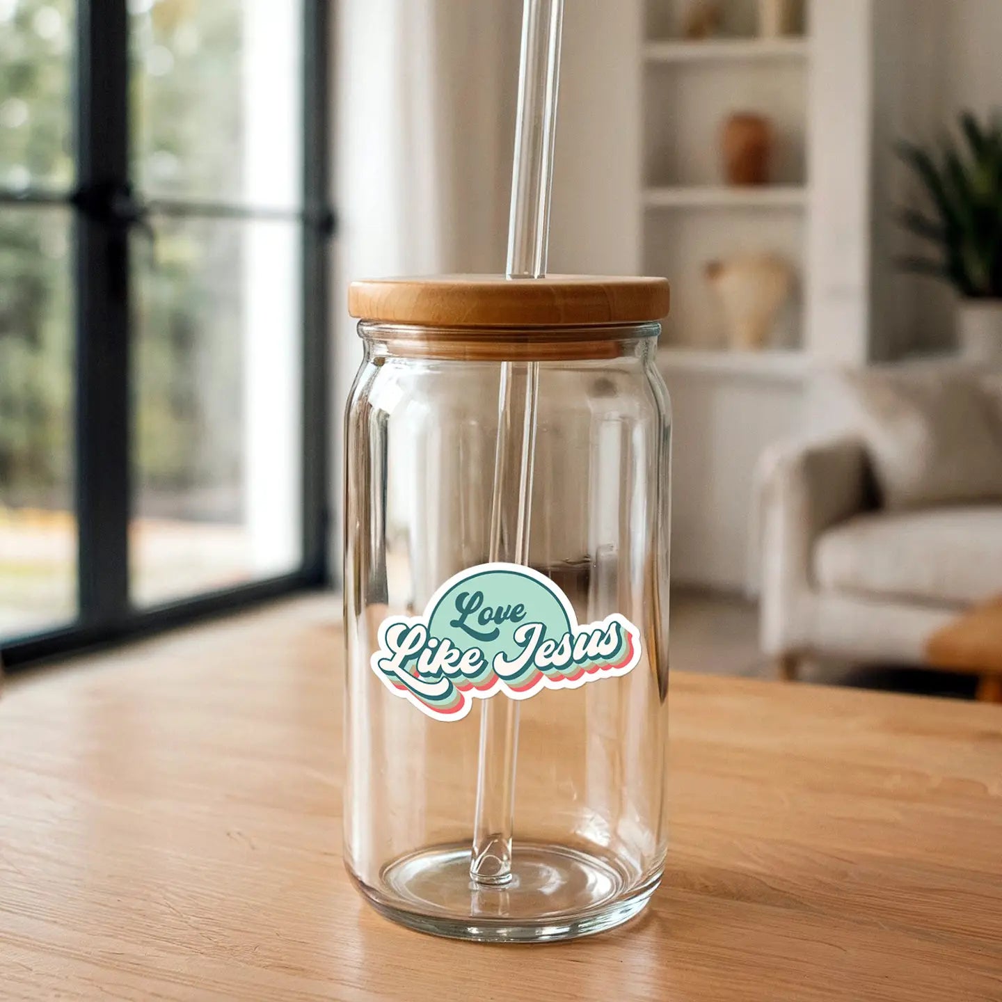 Clear glass tumbler with a bamboo lid, glass straw, and a teal, pink, and red "Love Like Jesus" sticker, placed on a light wooden table in a modern home with a blurred window and bookshelf