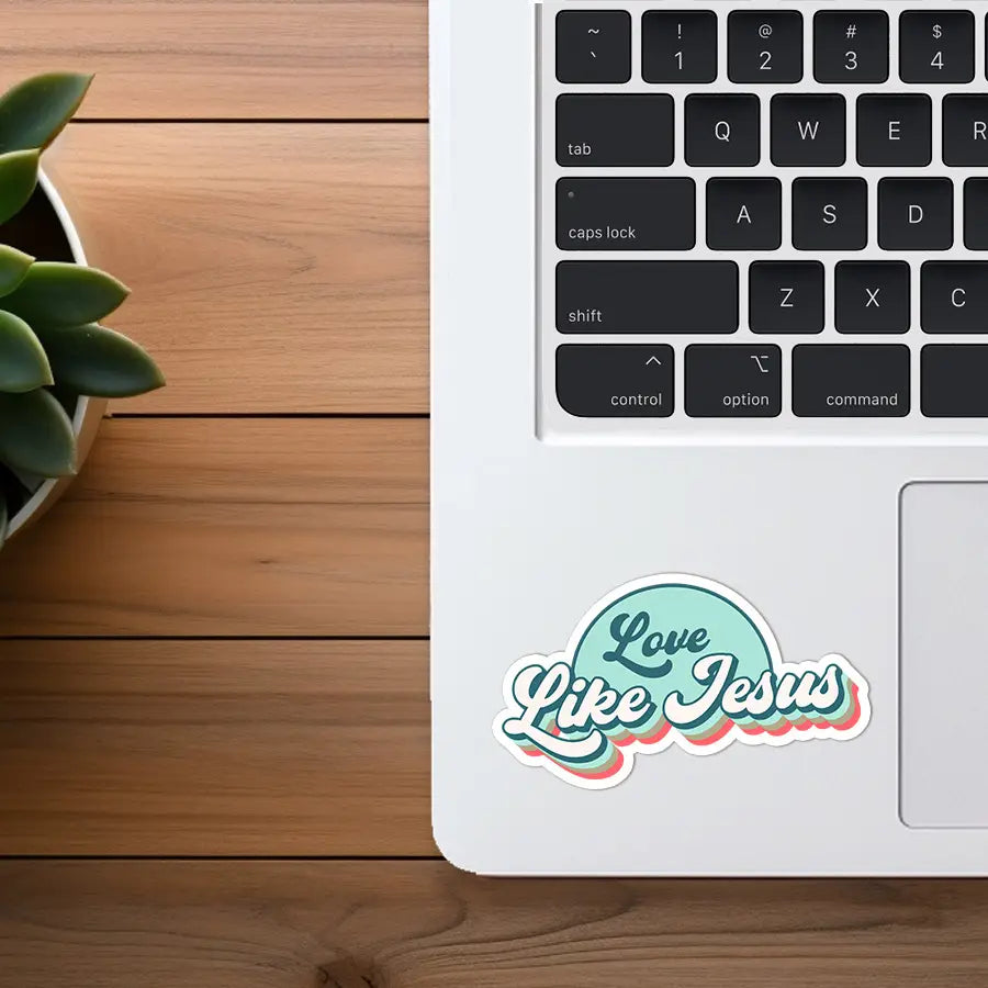 Photograph of a white Apple MacBook laptop on a wooden desk displaying an aqua green, white, and coral "Love Like Jesus" sticker with a green succulent plant visible on the left