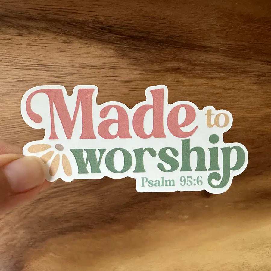 Sticker featuring the phrase Made to worship Psalm 95:6 with pink and green script lettering and a light brown flower graphic replacing the 'o' in worship, held by a hand against a warm wooden background