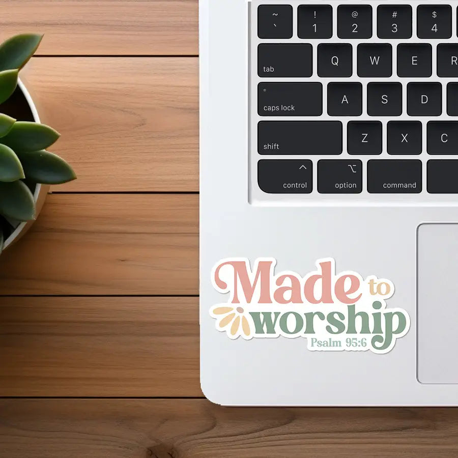 Sticker on laptop featuring the text Made to Worship Psalm 95:6 in pink green and beige retro fonts with a small daisy design on a white MacBook-style laptop keyboard next to a green succulent plant in a white pot on a warm wooden desk