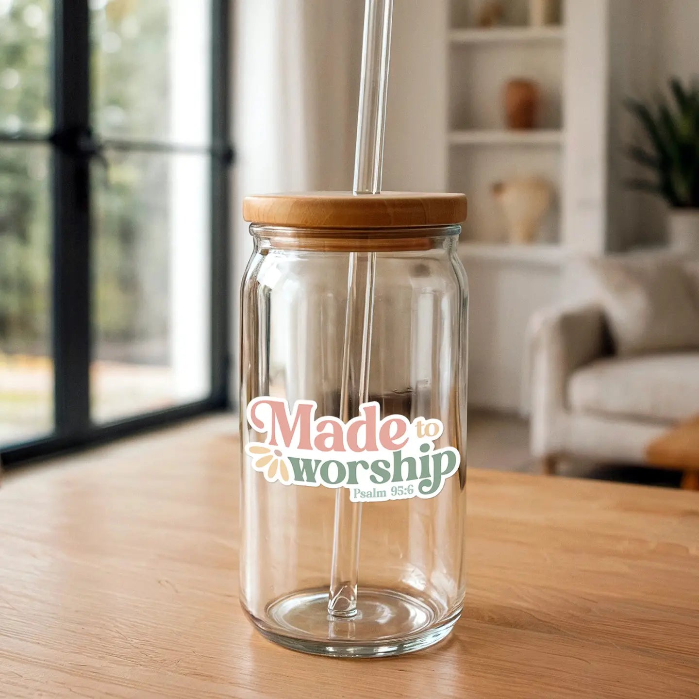 A clear glass tumbler with a bamboo lid and glass straw featuring a colorful "Made to worship Psalm 95:6" sticker resting on a light wooden table indoors with a blurred window and white bookshelf in the background