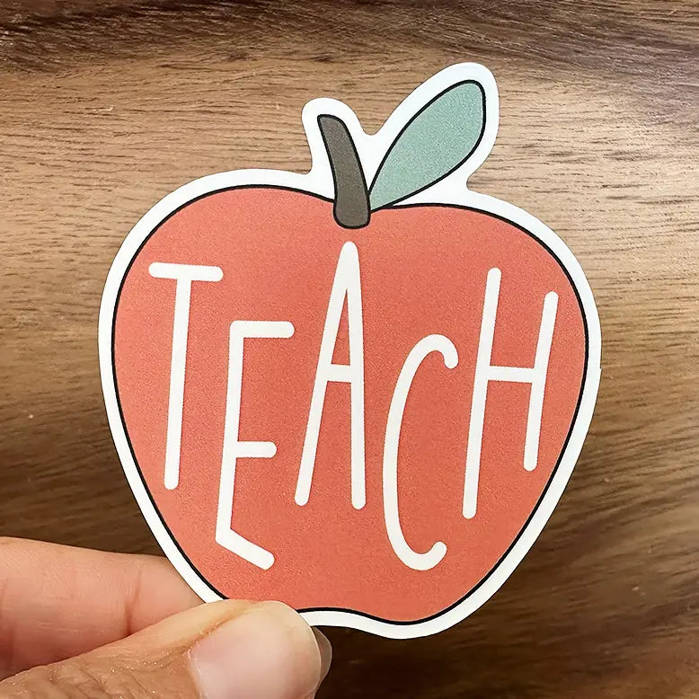 A hand holds a vibrant red apple-shaped sticker with a green leaf and brown stem, featuring the bold white word TEACH, against a warm wood grain background