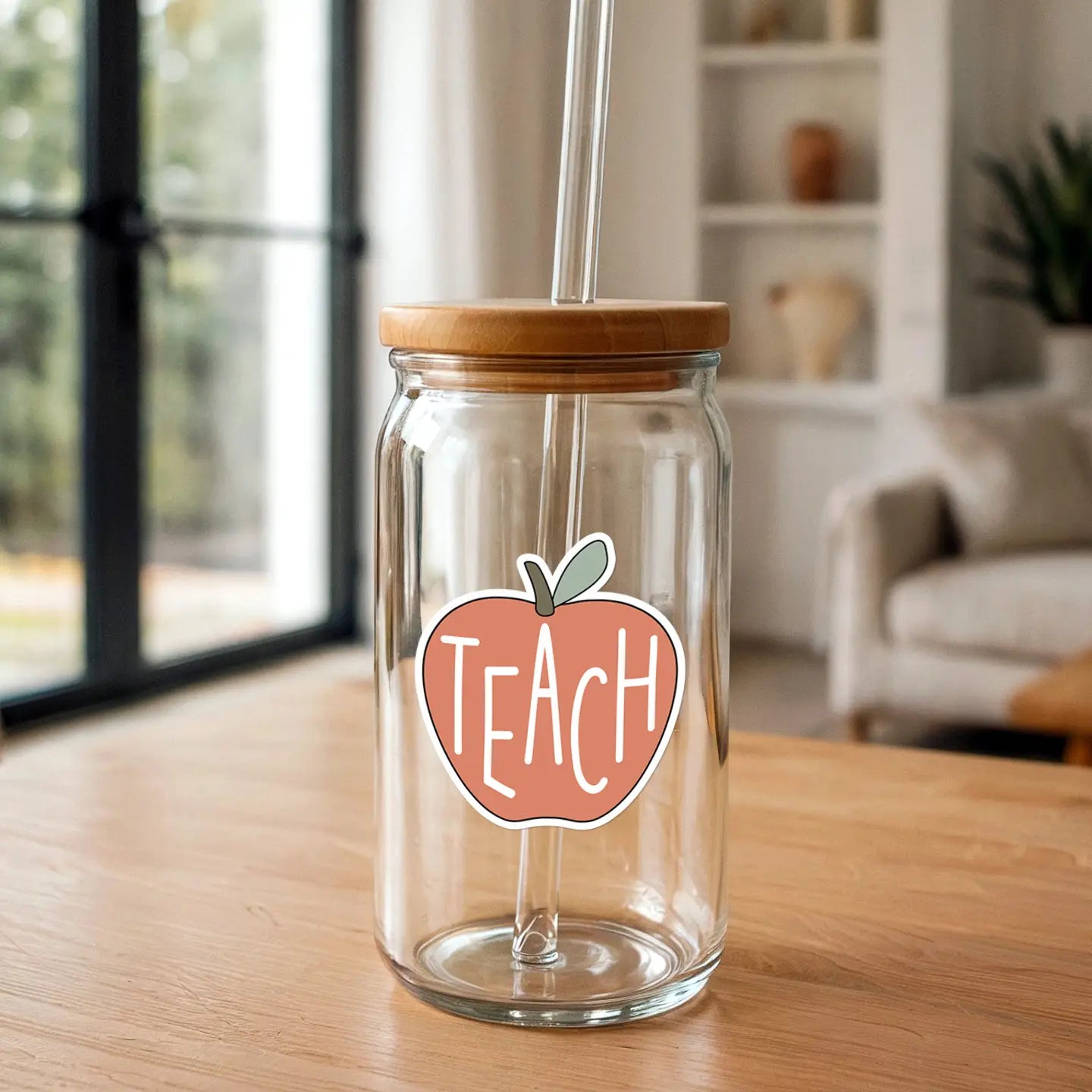 Photograph of a clear glass jar with a bamboo lid and glass straw featuring a peach-colored apple sticker with white TEACH text on a light wooden table with a blurred bright window and modern home interior in the background