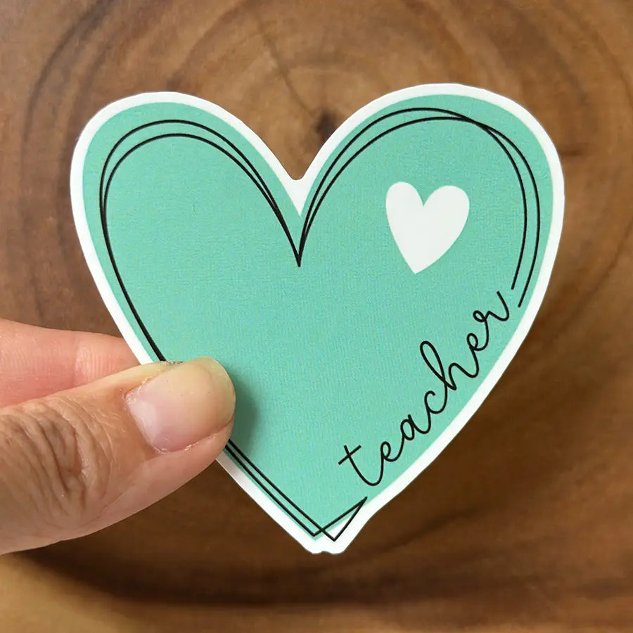 Sticker featuring a teal heart with a decorative black outline, a small white heart, and the word 'teacher' in script font, held by fingers against a warm wood grain surface