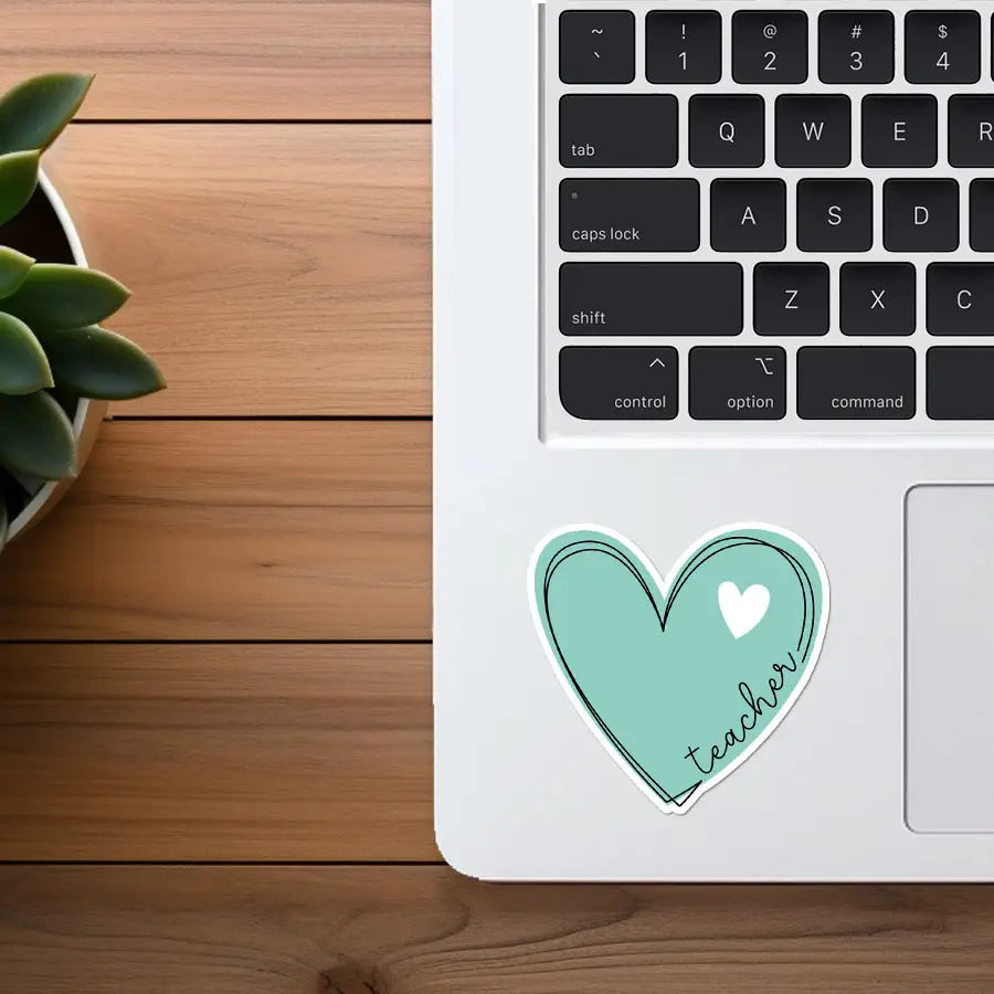A teal heart-shaped teacher sticker with a small white heart and black cursive text affixed to a white MacBook keyboard beside a green succulent plant on a wooden desk