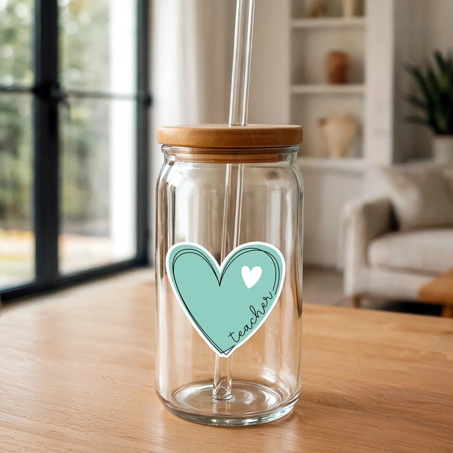 Clear glass jar with bamboo lid and glass straw featuring a turquoise heart sticker with white heart and "teacher" text on a wooden table with a blurred modern living room background