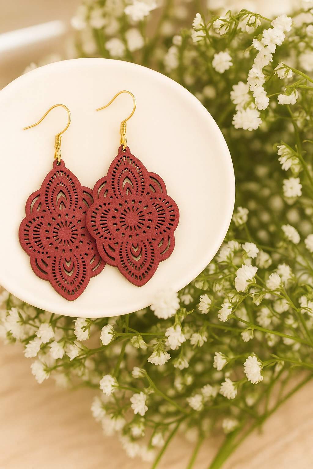 Photograph of a pair of maroon laser-cut earrings featuring intricate mandala patterns and gold hooks, displayed on a white dish with a soft background of white baby's breath flowers