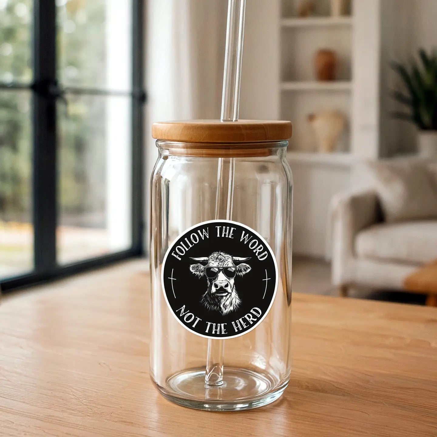 A clear glass tumbler with a bamboo lid and glass straw prominently displaying a circular black and white sticker of a cool cow in sunglasses with the text 'FOLLOW THE WORD NOT THE HERD' on a wooden table inside a bright room with a window and a blurred bookshelf