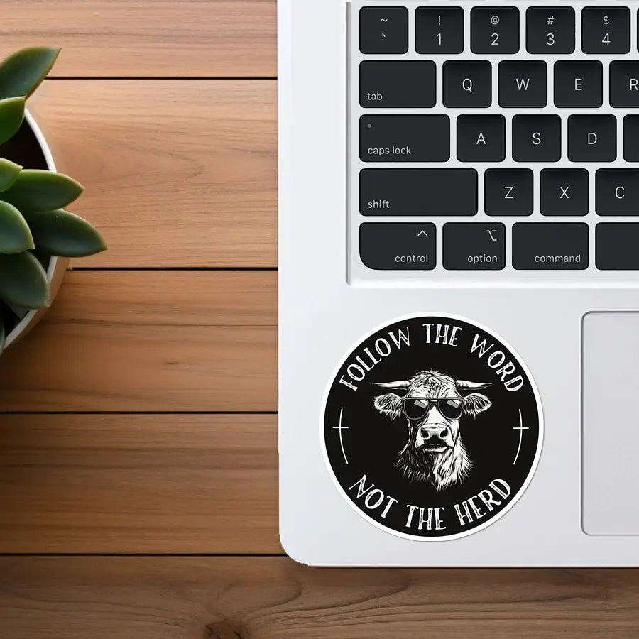 A black and white circular sticker featuring a cool cow in sunglasses and the phrase FOLLOW THE WORD NOT THE HERD on a white laptop with a green succulent plant and wooden desk in the background