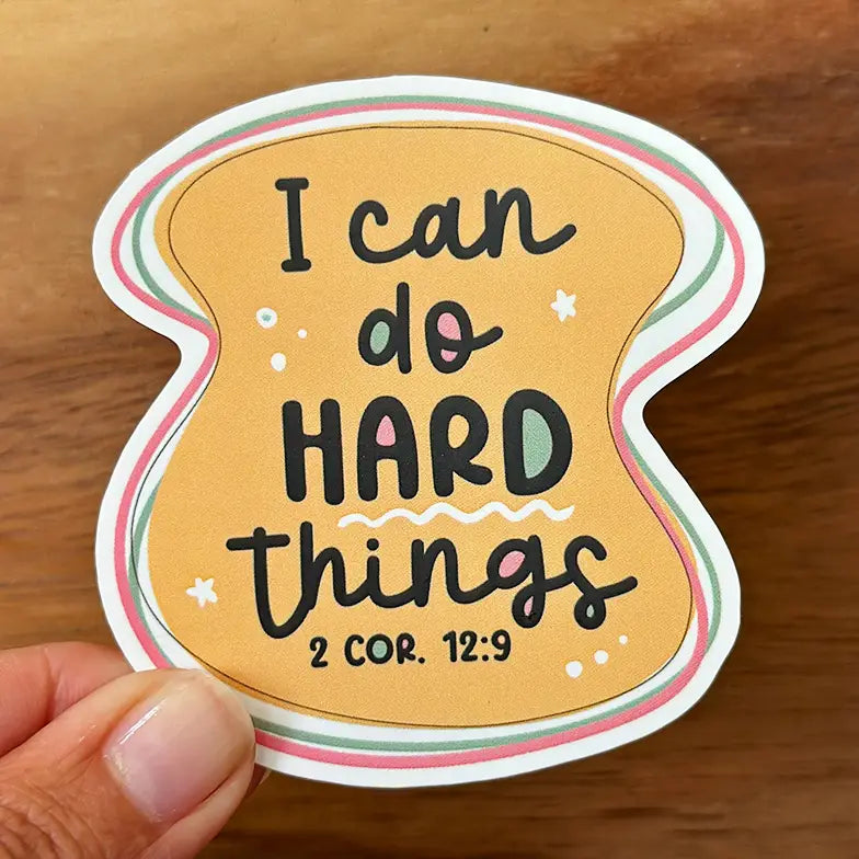 Sticker featuring the quote I can do HARD things 2 COR 12:9 in black and teal lettering on an orange toast-shaped background with pink and green striped borders held by a hand against a wooden surface