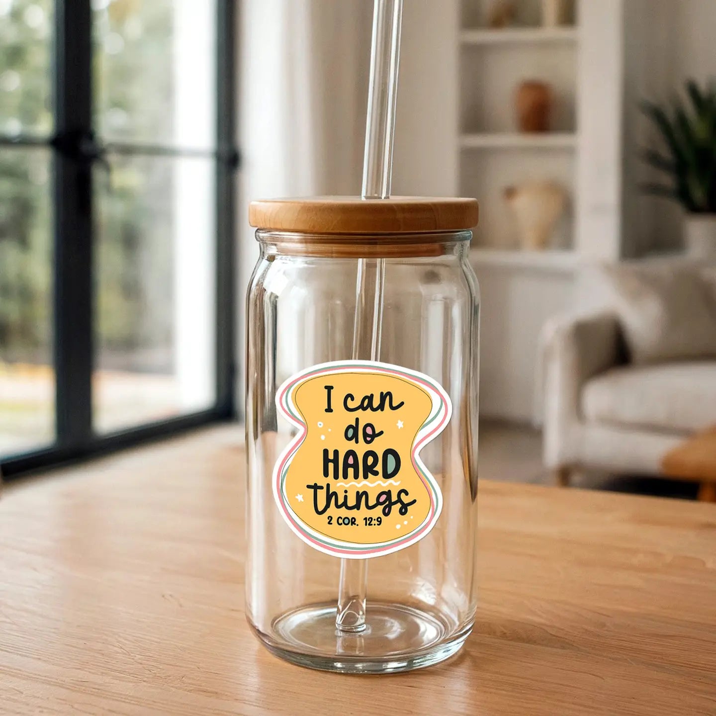 Clear glass jar with a yellow sticker reading I can do HARD things 2 COR 12:9, featuring a wooden lid and a clear glass straw, positioned on a light wooden table in a bright indoor setting