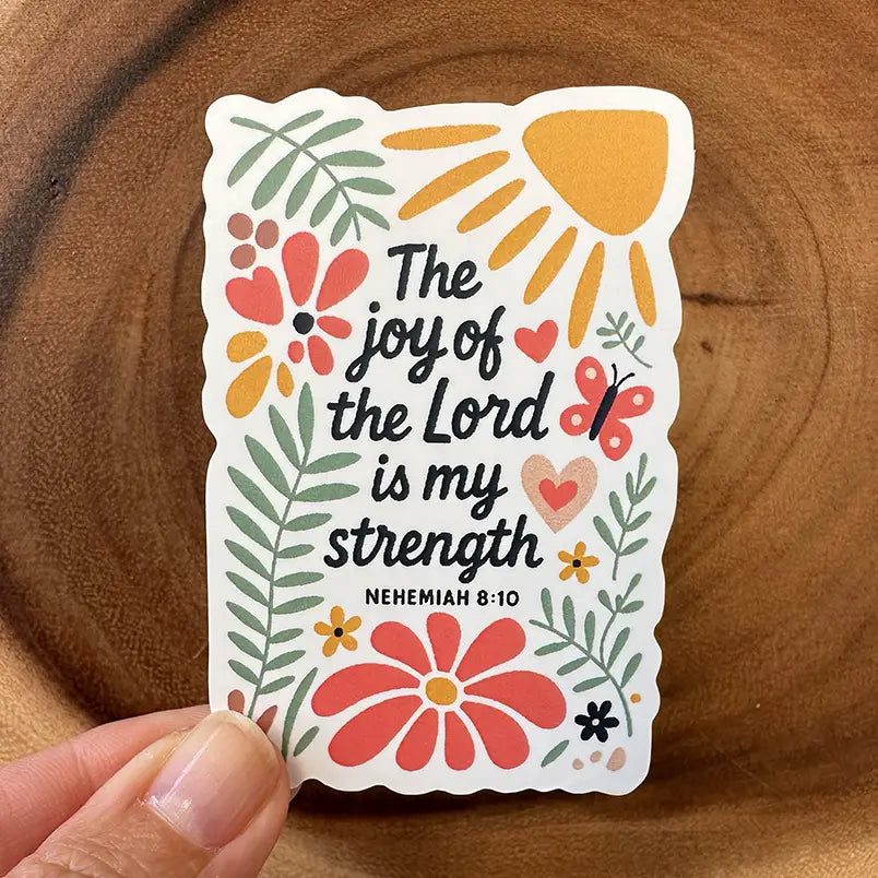 A vibrant sticker with the quote The joy of the Lord is my strength Nehemiah 8:10 adorned with a yellow sun, red and orange flowers, green leaves, and a pink butterfly, held by a hand against a wooden backdrop