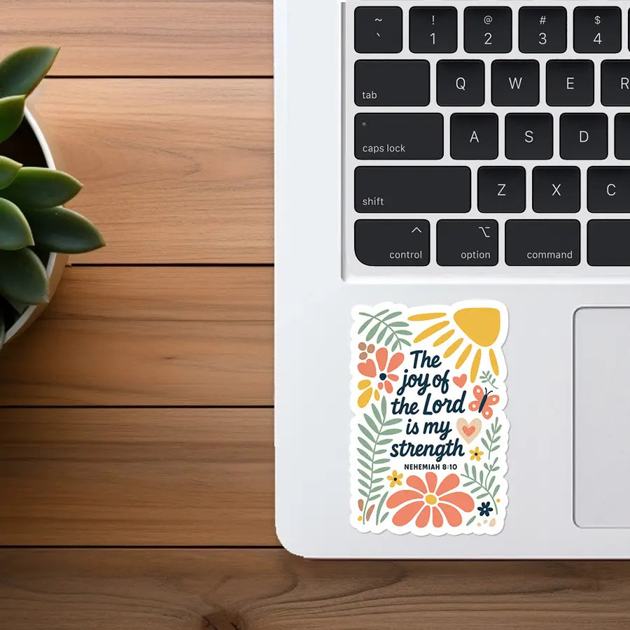 Photograph of a rectangular vinyl sticker on a silver laptop palm rest next to a green succulent, featuring the biblical quote "The joy of the Lord is my strength Nehemiah 8:10" with colorful illustrations of a yellow sun, orange flowers, and green foliage on a wooden desk