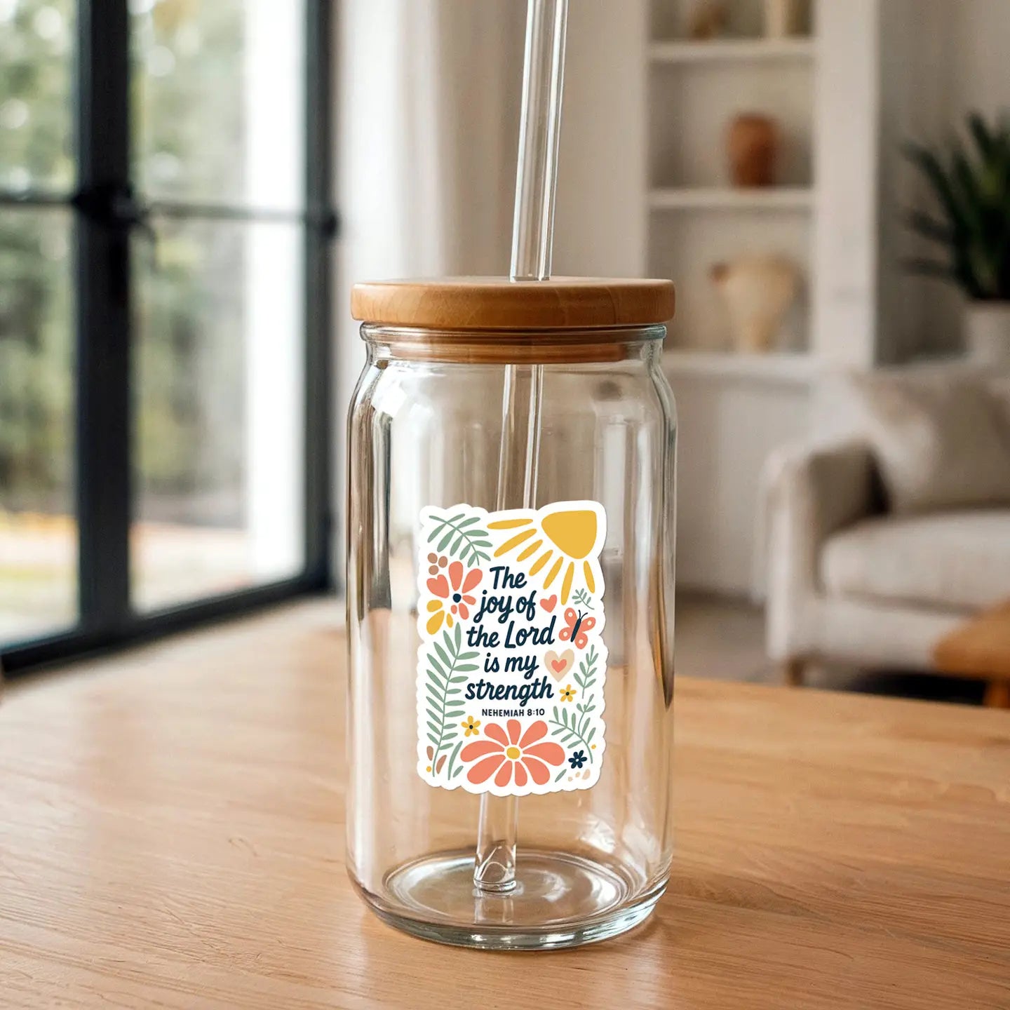 A clear glass can-shaped cup with a bamboo lid and glass straw features a colorful sticker depicting "The joy of the Lord is my strength" with orange flowers, green leaves, and a yellow sun, set on a light wooden table in a bright indoor room with a window