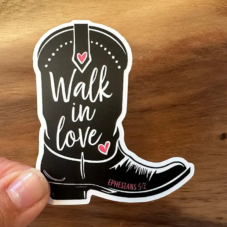 Sticker art of a black cowboy boot emblazoned with white script 'Walk in love', two vibrant pink hearts, and small pink text 'Ephesians 5:2', held by a hand against a warm wooden surface