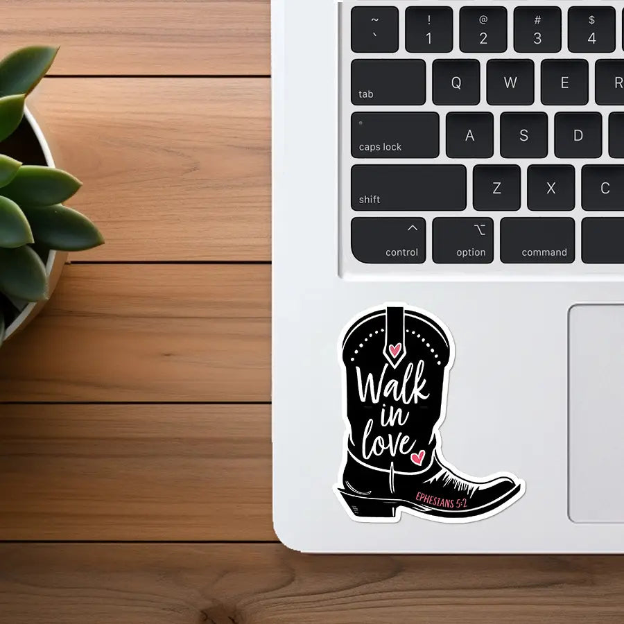 Photograph of a black cowboy boot sticker emblazoned with "Walk in love" in white script, pink heart details, and "EPHESIANS 5:2" displayed on a white laptop keyboard next to a wooden desk and a green succulent plant