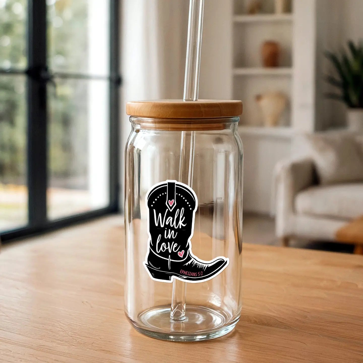 Clear glass jar with a bamboo lid and glass straw featuring a black cowboy boot sticker adorned with pink hearts and the text 'Walk in love Ephesians 5:2' on a light wooden table indoors