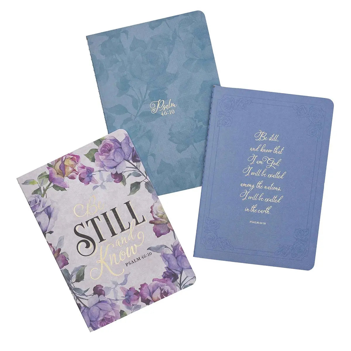 Three religious themed notebooks including a light blue one with a subtle floral pattern and Psalm 46:10, a darker blue one with the full Be still and know that I am God quote, and a white one with vibrant purple and green floral illustrations and Be Still and Know text