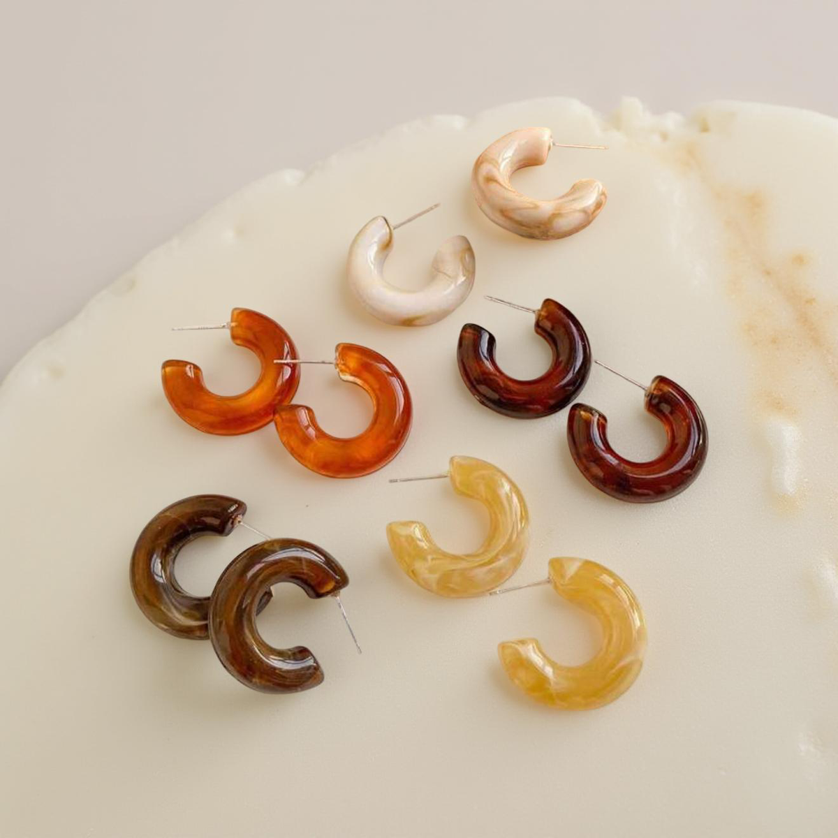 Multiple pairs of C-shaped resin hoop earrings in translucent amber, dark brown, creamy white, and pale yellow marbled patterns arranged on a light textured surface