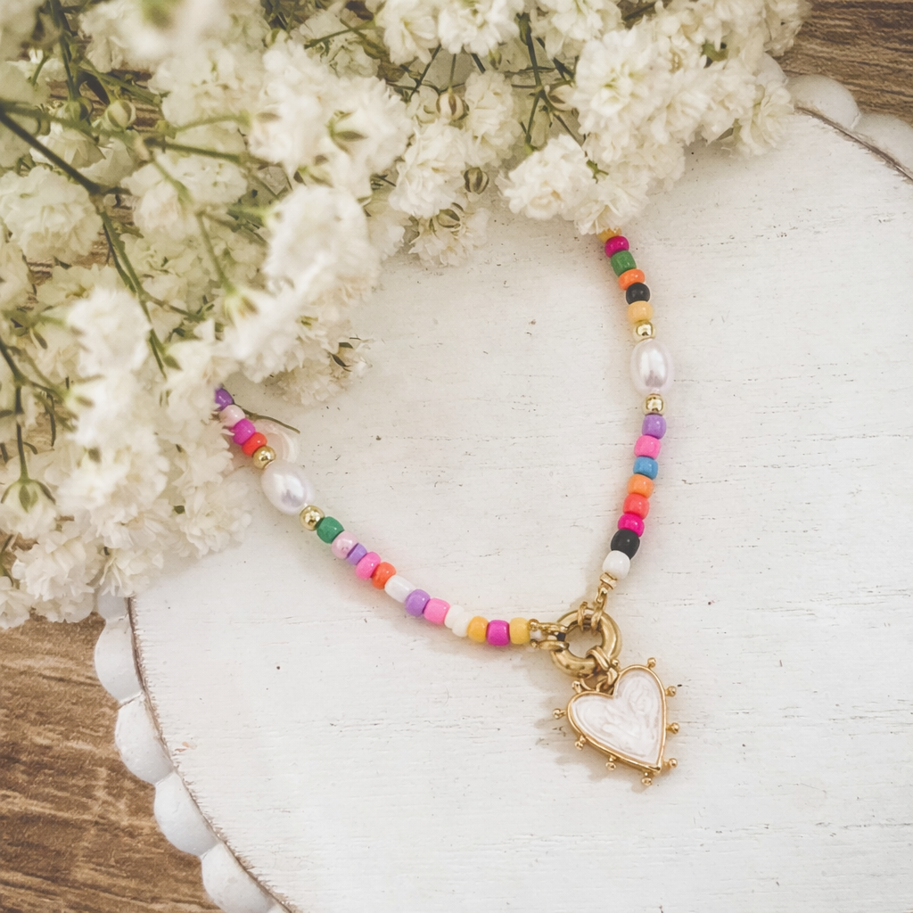 A vibrant beaded necklace featuring rainbow seed beads and baroque pearls with a unique white enamel heart pendant bordered by gold studs and a textured frame, displayed on a distressed white wooden surface next to delicate baby's breath flowers