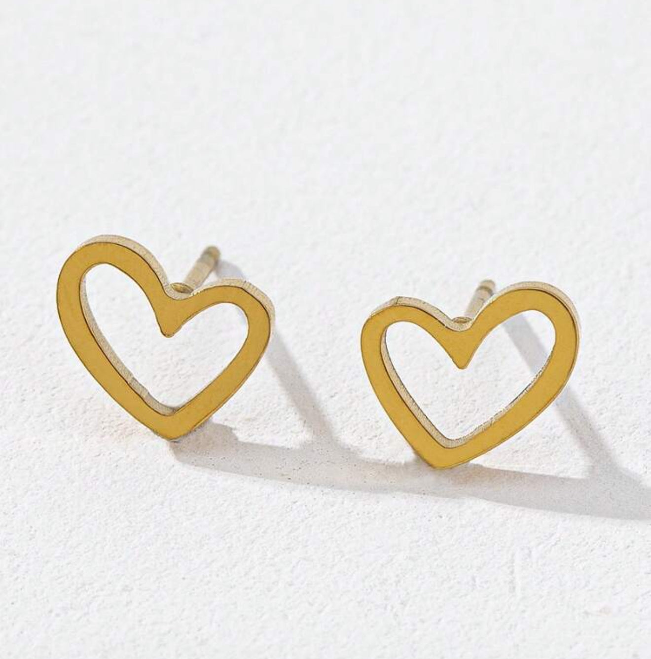 A pair of gold tone hollow heart outline stud earrings displayed on a clean white surface with subtle shadows