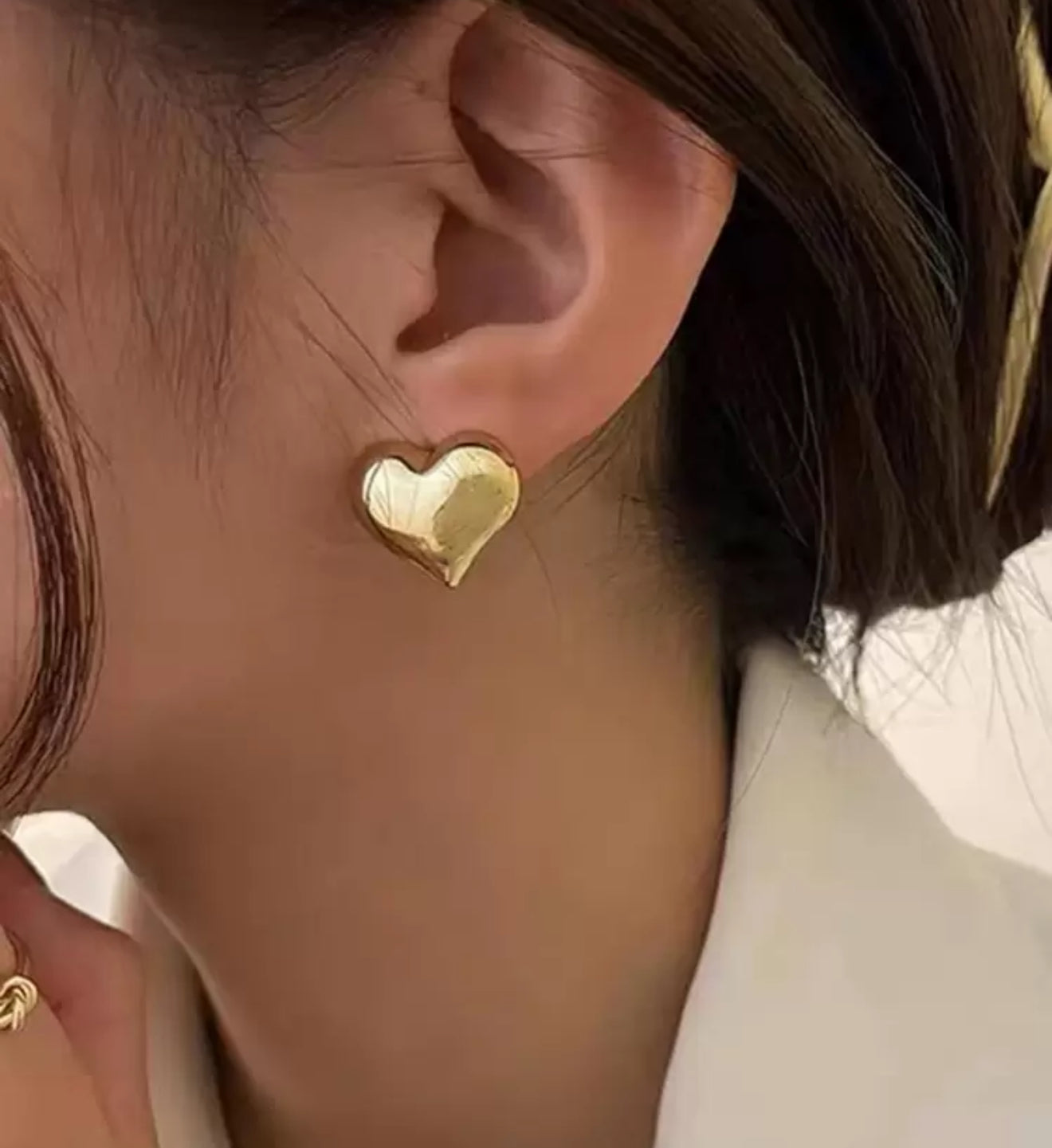 Close-up of a person's ear adorned with a shiny gold heart-shaped stud earring, dark hair, and a white garment