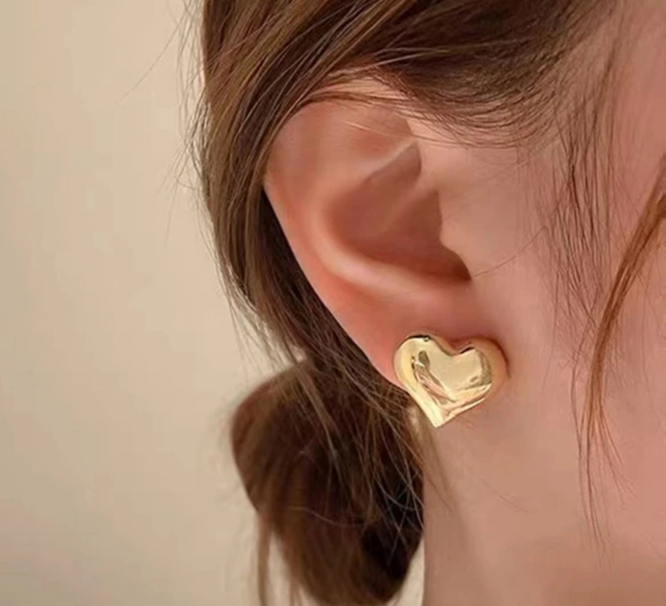 Photograph of a woman's ear adorned with a shiny gold puffy heart stud earring and visible light brown hair