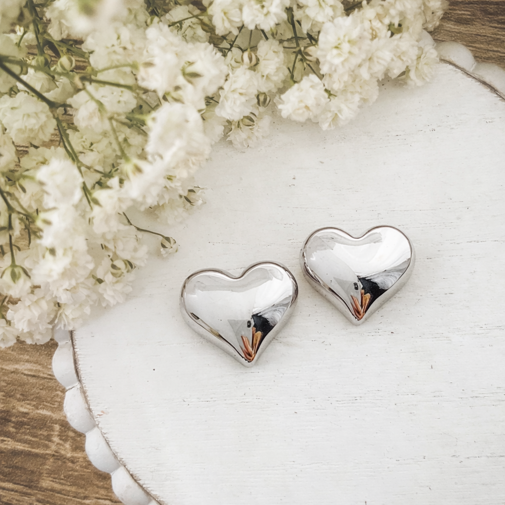 Close-up photograph featuring two reflective silver heart-shaped objects on a white distressed scalloped wooden stand, with a blurred arrangement of delicate white baby's breath flowers in the upper left corner
