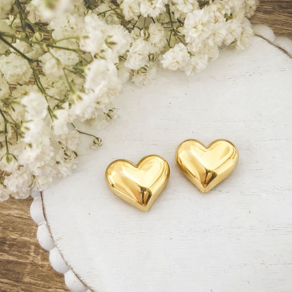 Digital Photograph of two polished golden heart objects resting on a distressed white wooden tray with a beaded edge, alongside delicate clusters of white baby's breath flowers on a soft focus wooden background