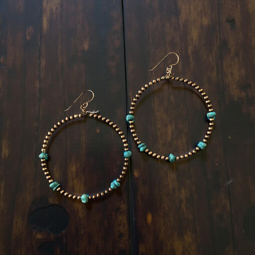 A pair of large beaded hoop earrings featuring small gold-toned beads and rough blue-green turquoise nuggets displayed on a dark rustic wooden surface