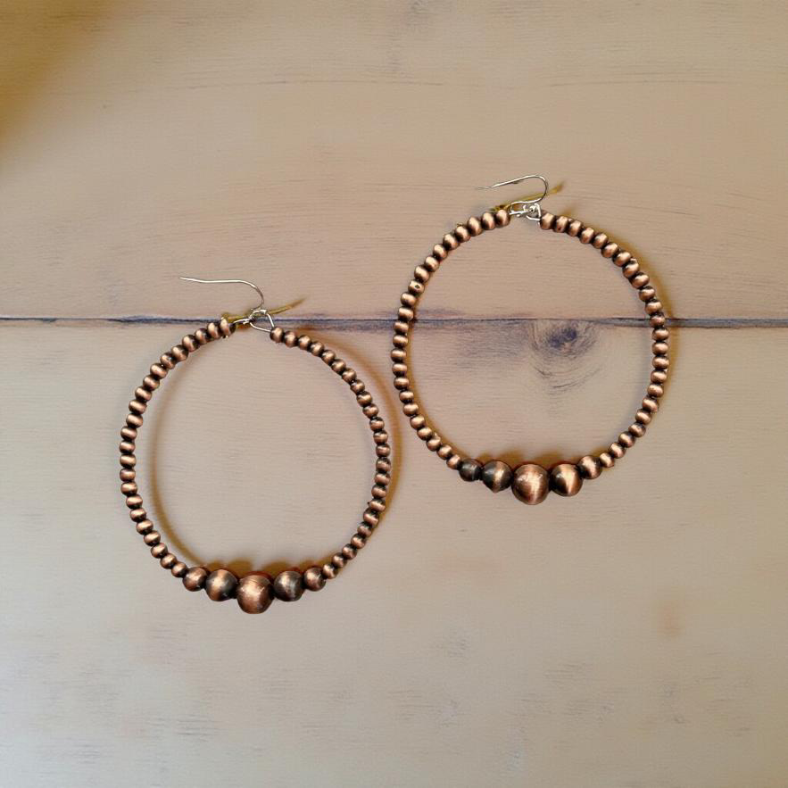 Jewelry a pair of shiny copper-tone beaded hoop earrings featuring three larger graduated accent beads at the bottom of each hoop presented on a light brown wooden surface