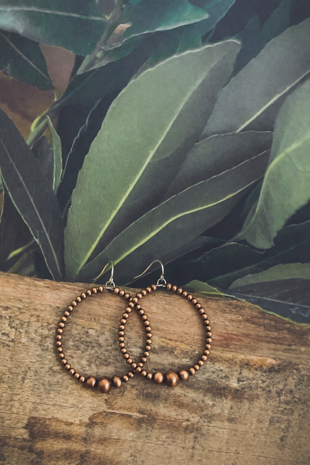 Photograph of glistening copper beaded hoop earrings with silver hooks and prominent accent beads, resting on textured rustic wood before a vibrant deep green leafy background