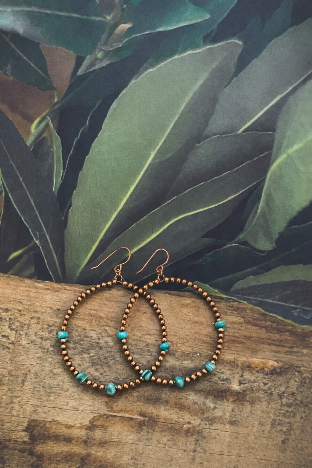Handmade copper bead hoop earrings featuring vibrant turquoise stones and rose gold hooks on a rustic wood plank with a dark green leafy background