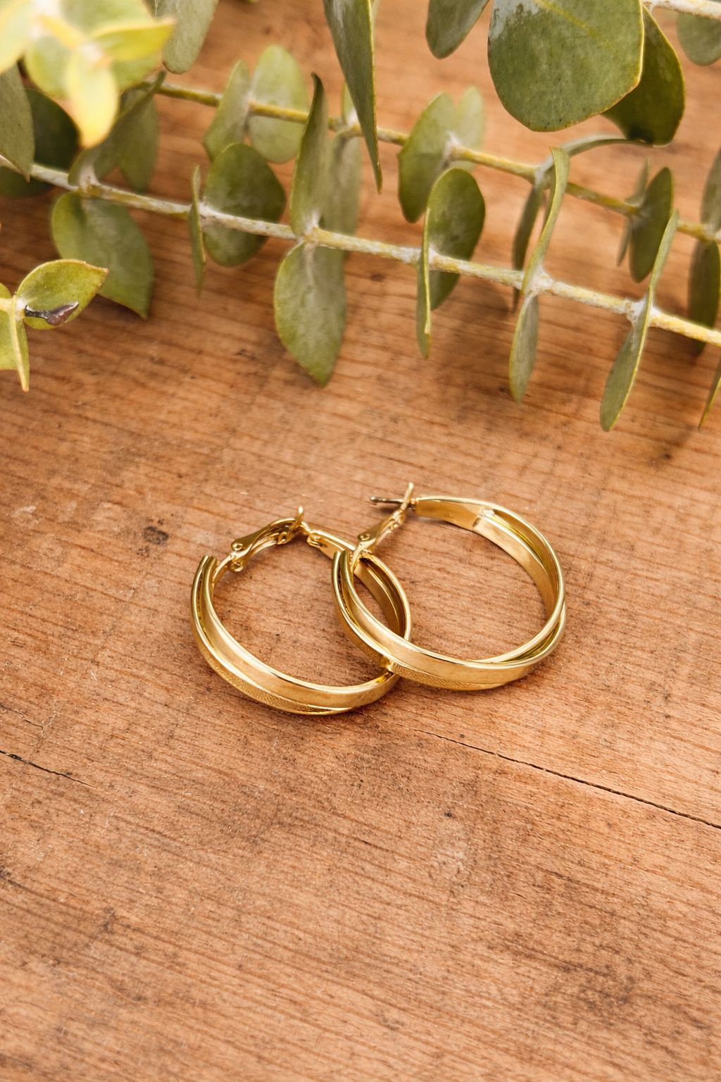 A pair of gold hoop earrings featuring mixed matte and shiny finishes with a textured pattern rests on a rustic wooden table beside a green eucalyptus branch