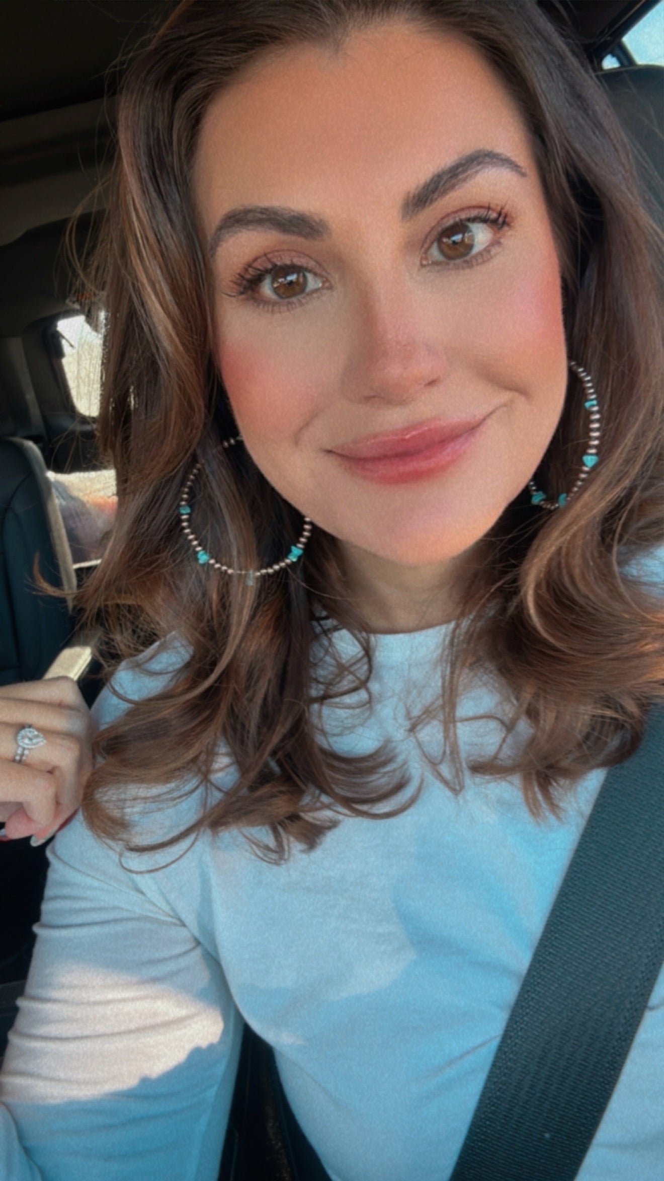Photograph of a smiling woman taking a car selfie with brown wavy hair, wearing large silver hoop earrings with turquoise beads and a diamond ring on her left hand